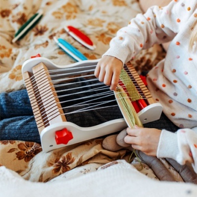 Criança a usar tear de madeira para tecer fios coloridos numa cama com manta castanha