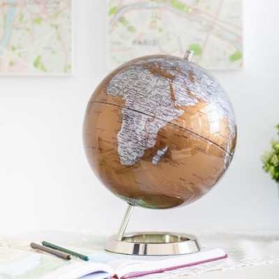 Globo terrestre decorativo em tons bronze e cinza numa mesa com caderno e mapas ao fundo