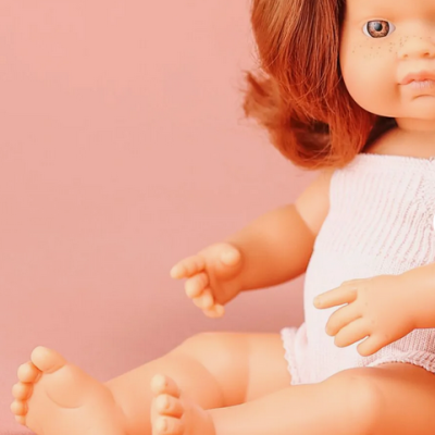 Boneca de bebé com cabelo castanho avermelhado e roupa branca sentada em fundo cor-de-rosa