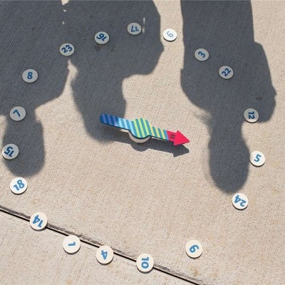 Relógio solar artesanal com ponteiro colorido e números em círculos brancos sobre cimento.