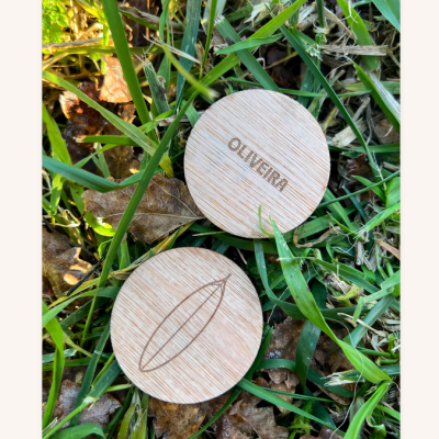 Dois objetos redondos de madeira clara numa superfície de relva e folhas, um com texto OLIVEIRA e outro com desenho gravado.