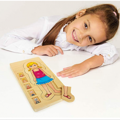 Menina sorridente junto a um quebra-cabeça de madeira de uma menina loira com vestido rosa e saia azul, numa mesa branca.