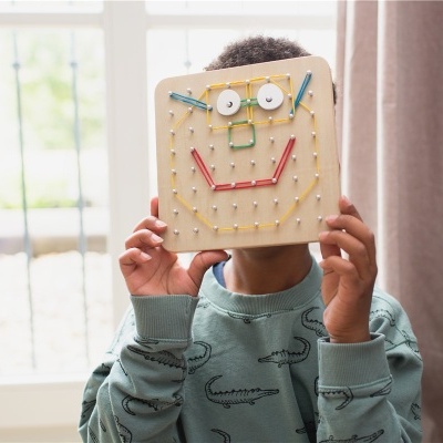 Menino segurando um quadro de madeira com pregos e elásticos coloridos formando uma cara sorridente