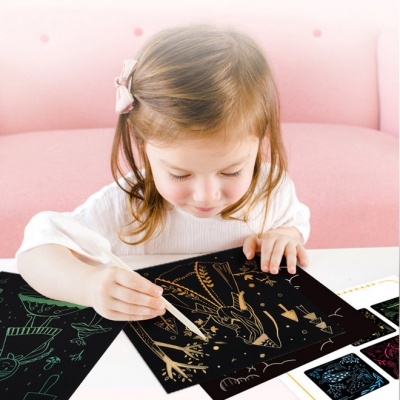 Criança a riscar desenhos dourados em papel preto numa mesa branca com sofá rosa ao fundo.