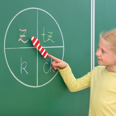 Criança a apontar para quadro verde com círculo dividido em quatro partes e letras z, tz, k e ck.