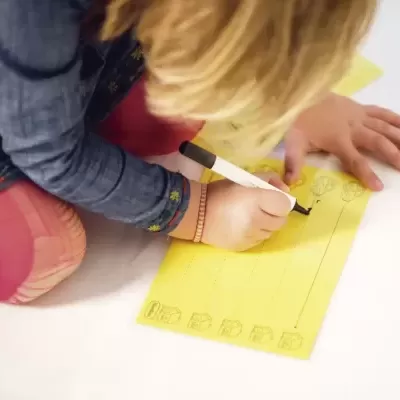 Criança desenha numa folha amarela com caneta preta.