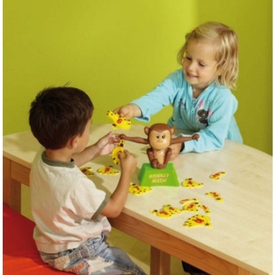 Crianças a jogar jogo de macaco com peças amarelas e base verde numa mesa de madeira clara.