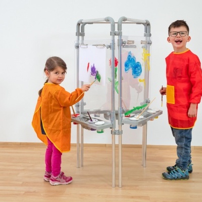 Duas crianças a pintar num cavalete transparente de quatro lados com bandejas e tintas.