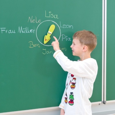 Menino a apontar para boneco amarelo num quadro verde com nomes escritos a giz.