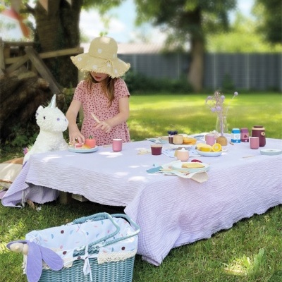 Mesa de piquenique com toalha lilás, pratos e copos, criança com vestido rosa e chapéu, boneco de pelúcia lhama, cesto azul no relvado