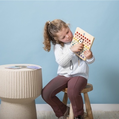 Menina a jogar com tabuleiro de peças coloridas sentada num banco de madeira junto a mesa bege