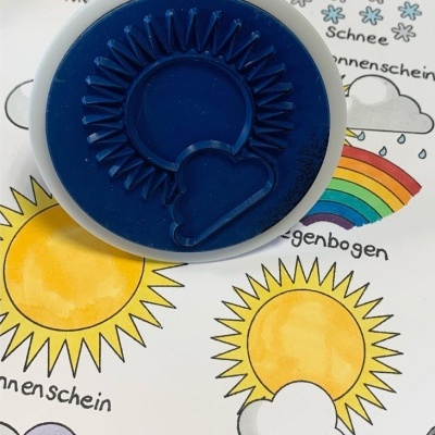 Carimbo de sol e nuvem sobre papel com desenhos coloridos de sol, nuvem e arco-íris e texto em alemão