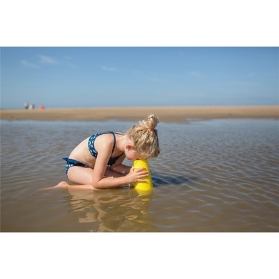 Menina com fato de banho azul sentada na praia junto à água, segurando um objeto amarelo.