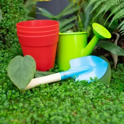 Conjunto de jardinagem com regador verde-limão, vasos vermelhos e pá azul em plantas verdes