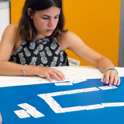 Jovem a jogar dominó com peças numéricas numa mesa branca e azul.