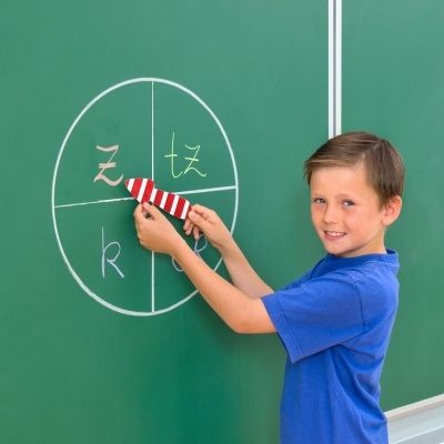 Menino com camiseta azul aponta com ponteiro vermelho e branco para quadro verde com círculo e letras coloridas.