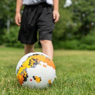 Bola de futebol branca com detalhes amarelos, laranja e preto numa relva verde com criança ao fundo.