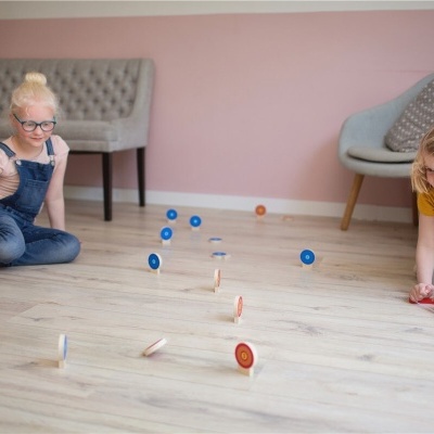 Duas crianças a jogar com piões coloridos no chão de madeira clara num quarto interior com parede cor-de-rosa.