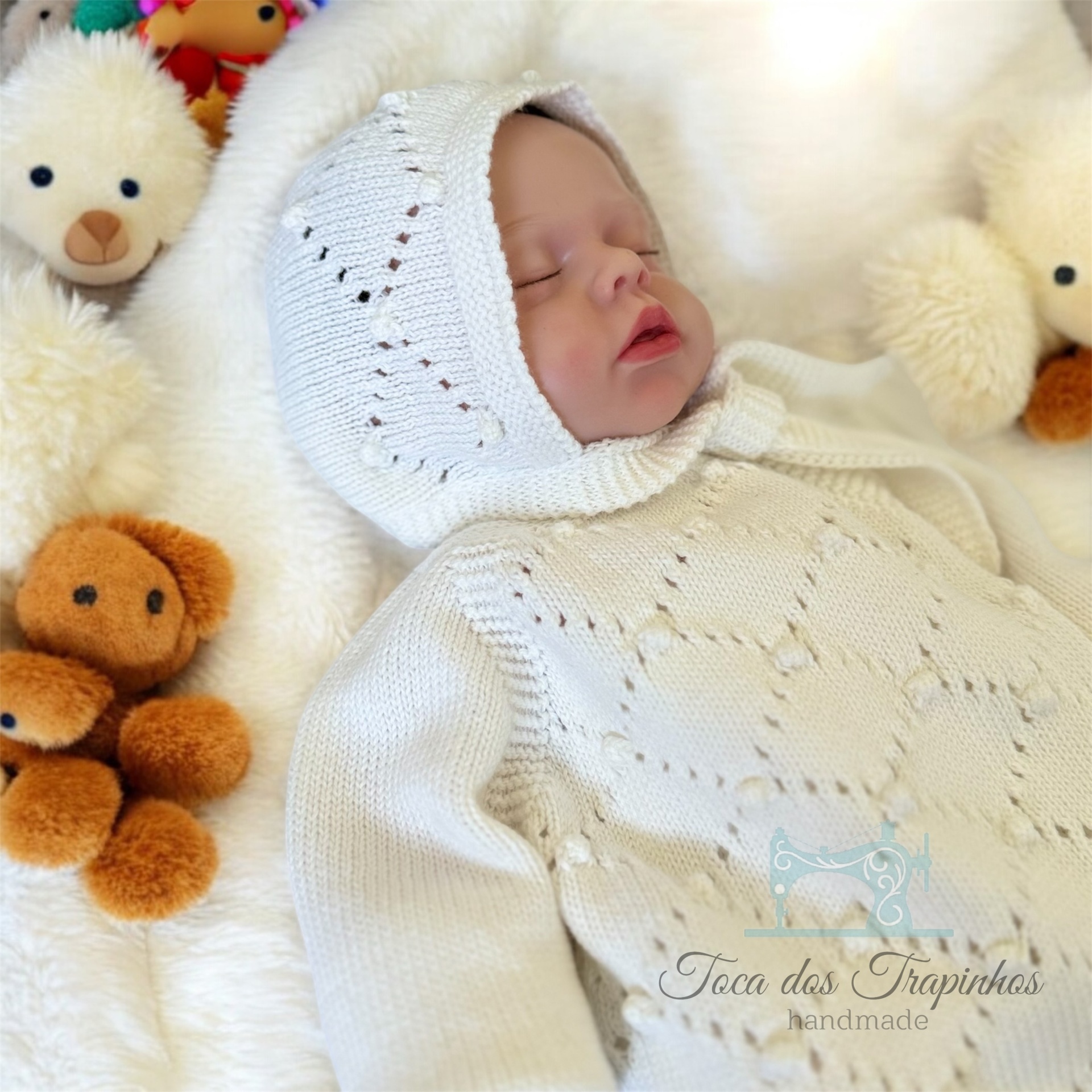 Bebé vestido com roupa de malha branca de capuz, rodeado por peluches sobre tecido branco