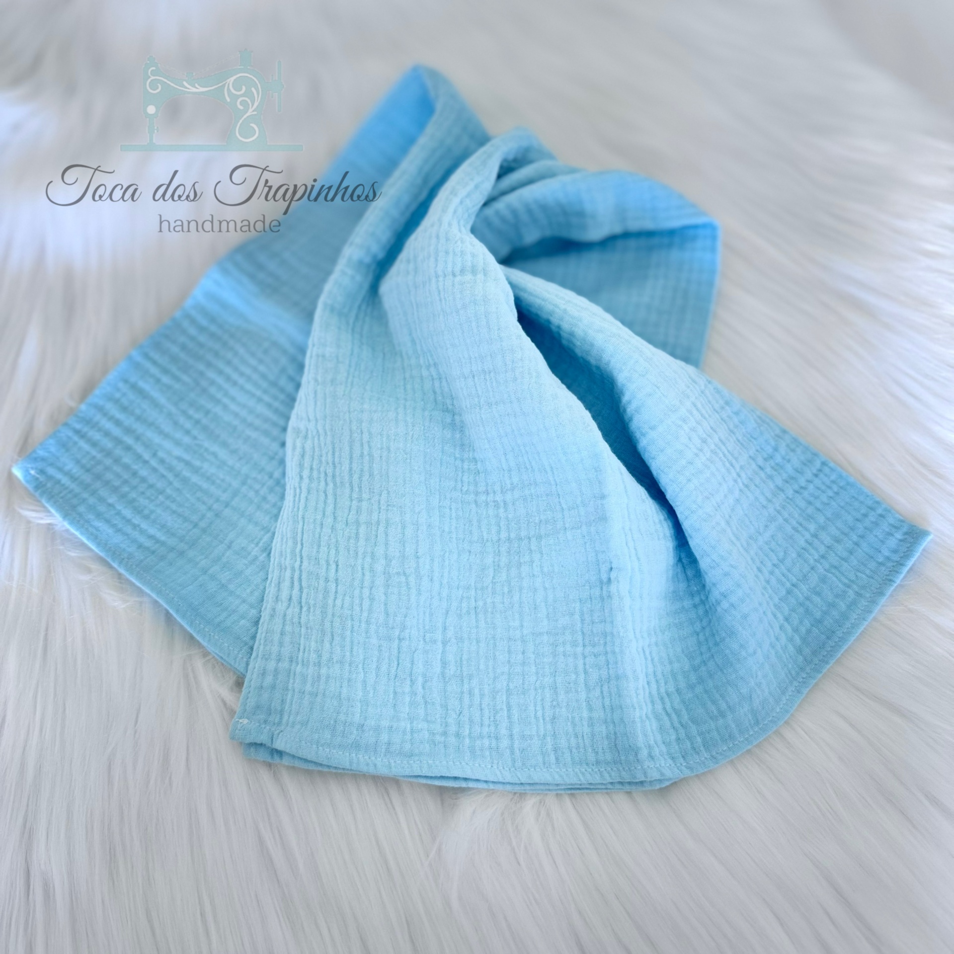 Lenço azul claro de tecido macio sobre fundo branco felpudo com texto da marca