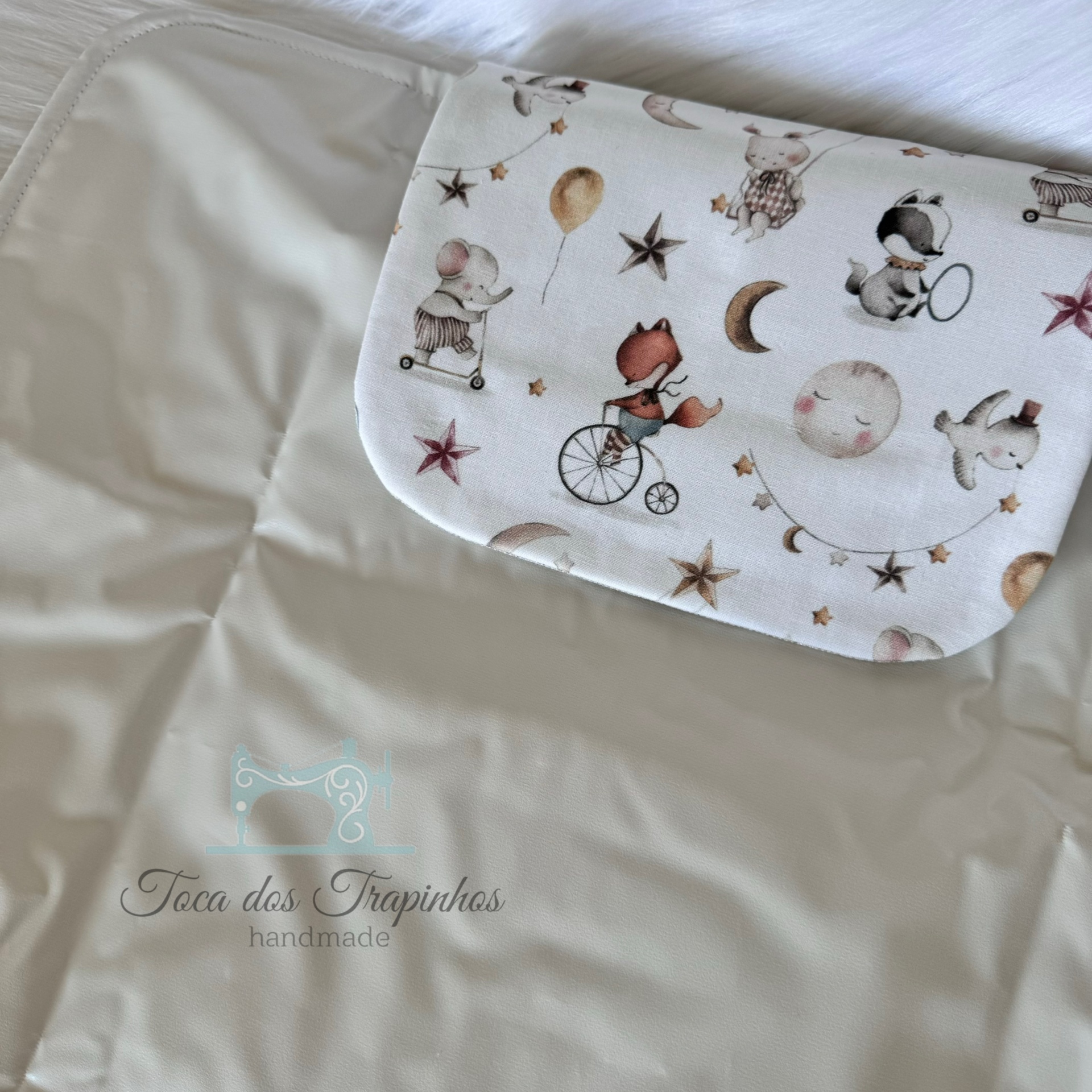 Tapete impermeável cinzento com bolso estampado com animais e estrelas, texto 'Toca dos Trapinhos handmade'.