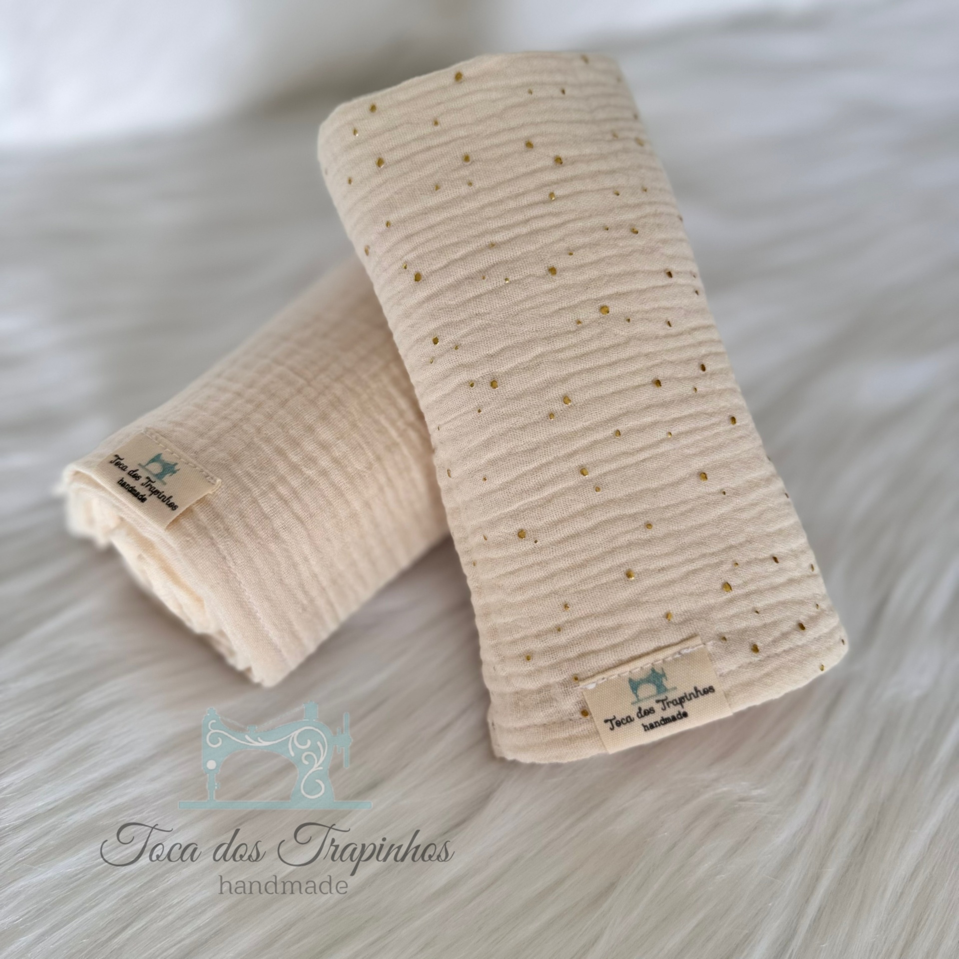 Dois panos creme com etiqueta Toca dos Trapinhos em superfície peluda branca