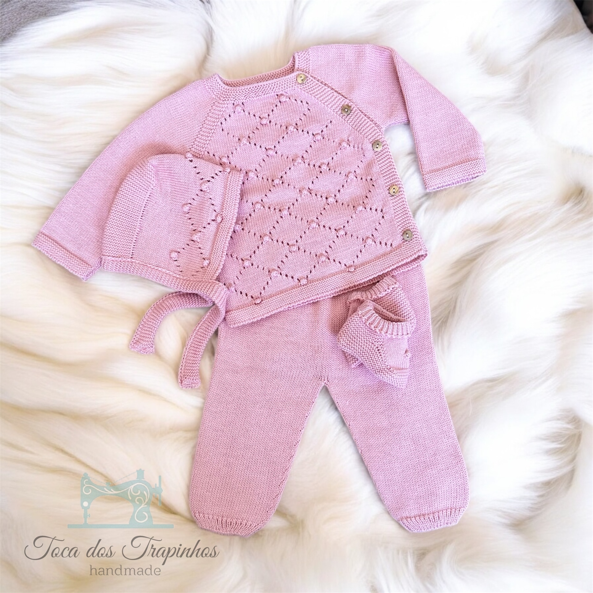 Conjunto de malha rosa para bebé com casaco, calças, gorro e pequena bolsa, sobre fundo branco felpudo