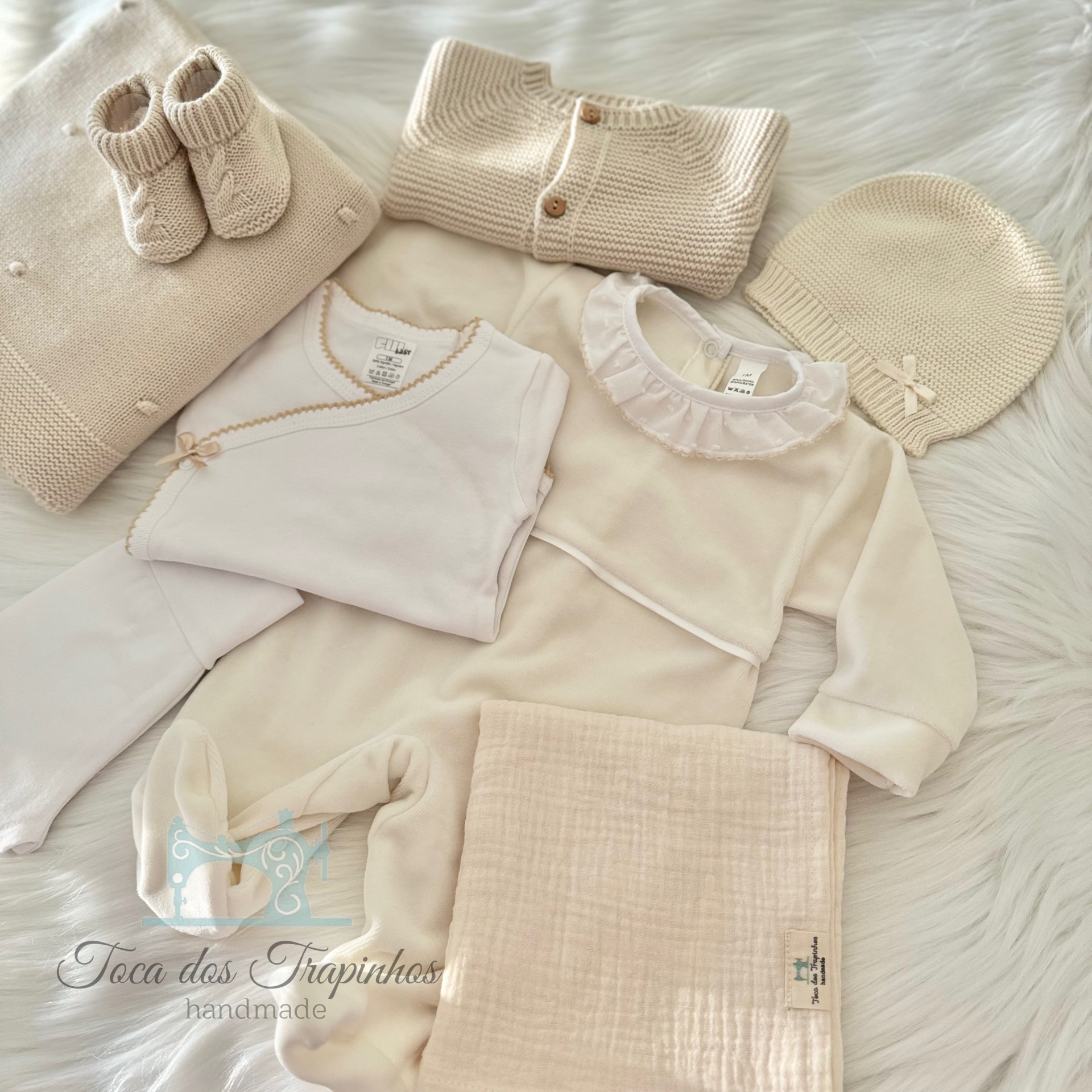 Conjunto creme de roupa de bebé com botinhas, casaco, touca, body e manta sobre superfície branca peluda