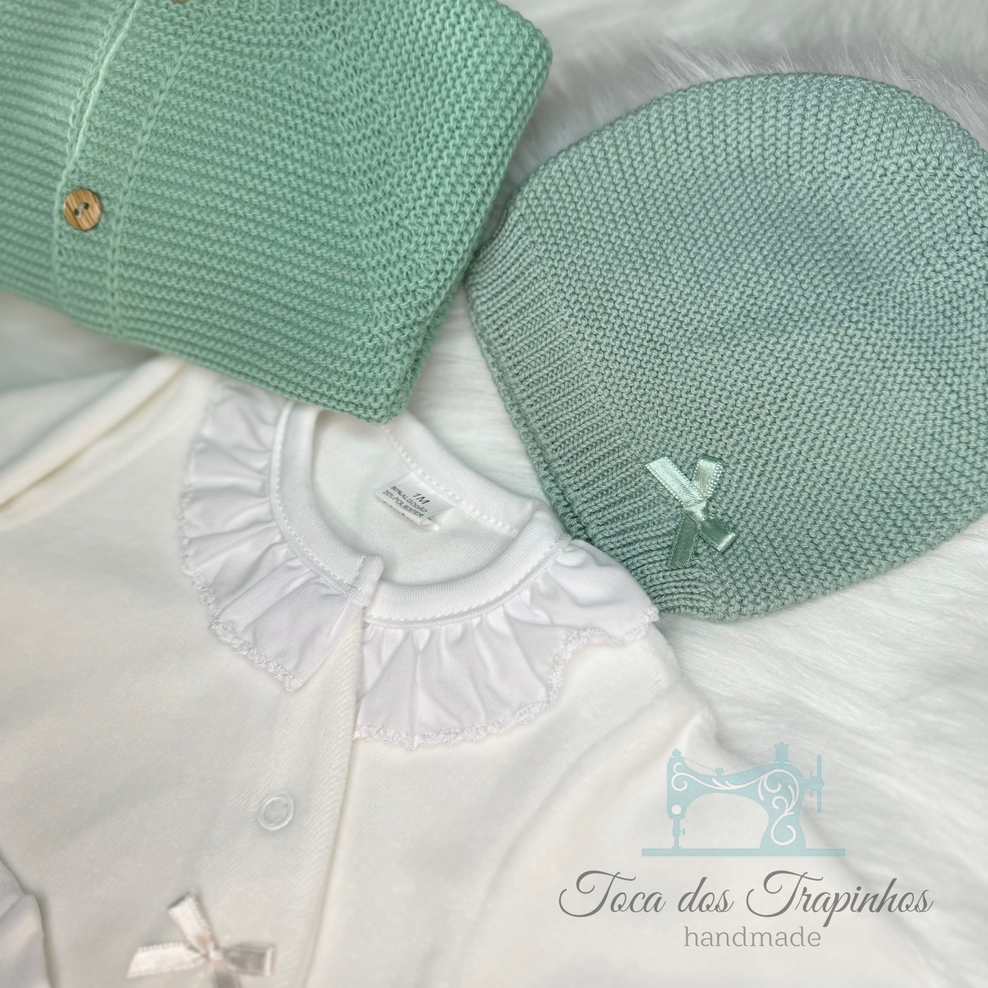 Conjunto para bebé com casaco branco de gola franzida, gorro e casaco verde água em tricô, com botão de madeira.