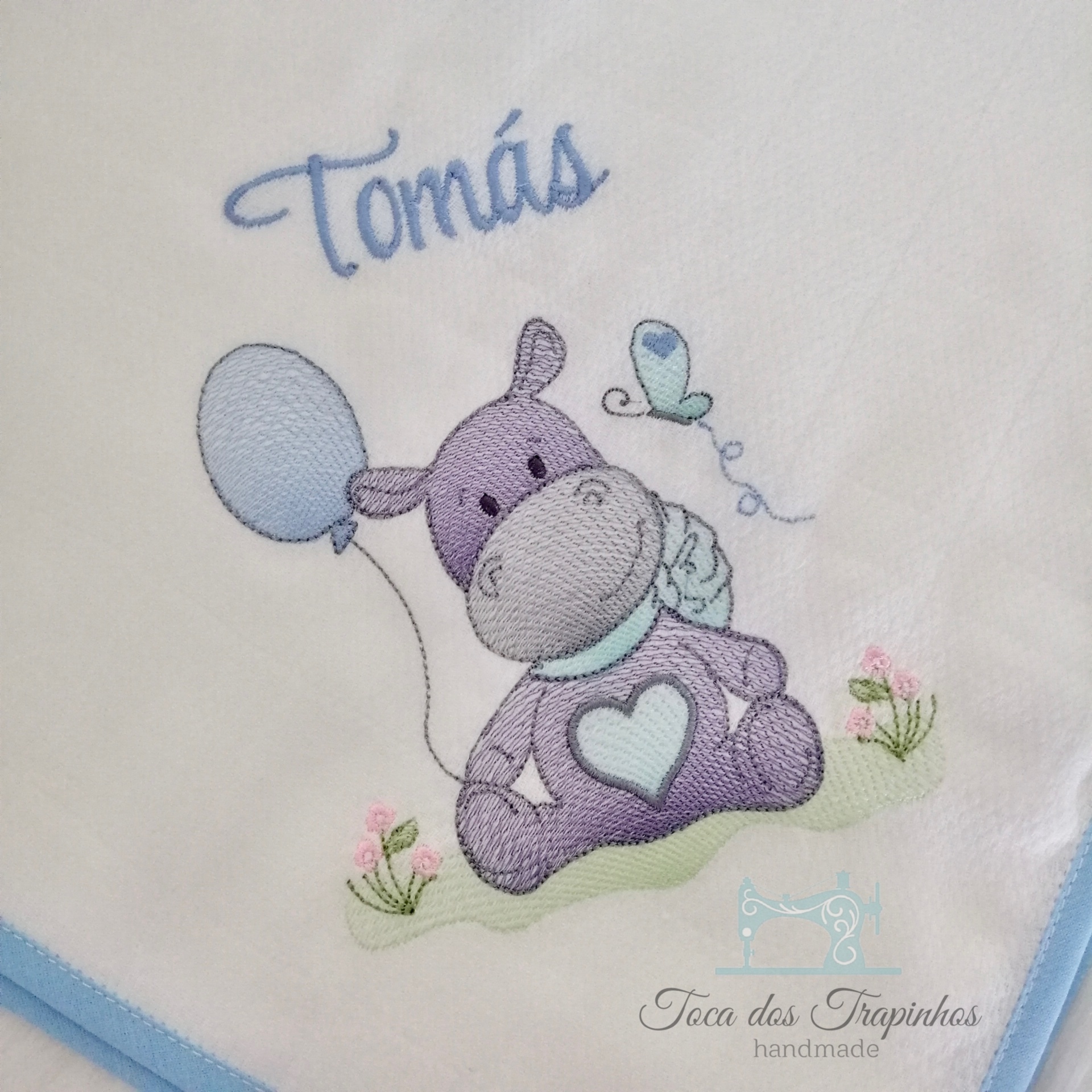 Fralda branca com bordado de hipopótamo lilás e balões azuis com o nome Tomás