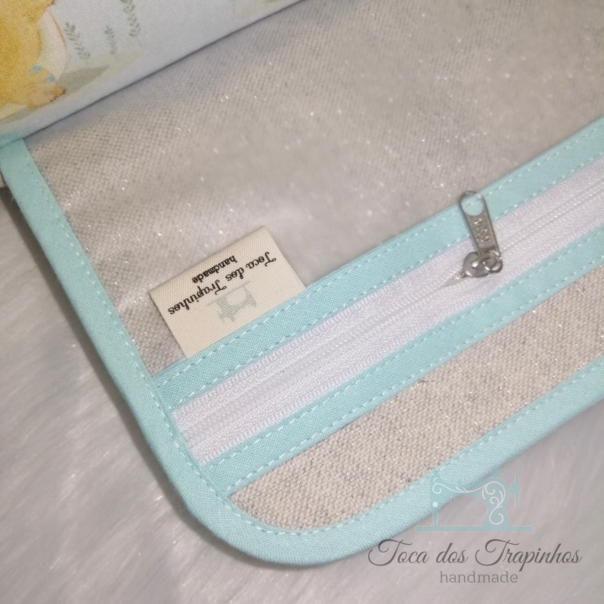 Estojo bege com fecho branco e borda azul clara, etiqueta 'Toca dos Trapos handmade'