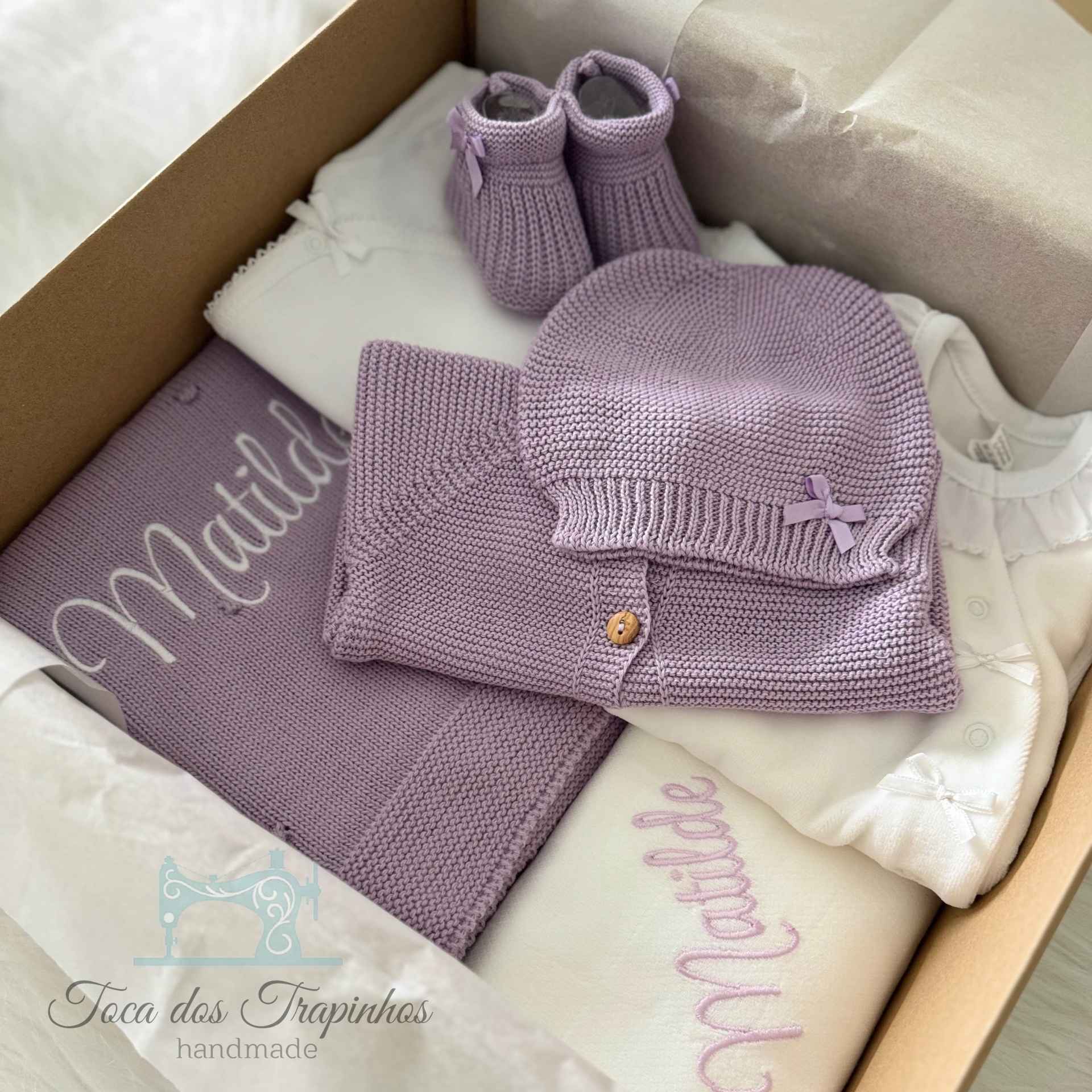 Conjunto de roupa de bebé lilás e branco numa caixa com manta bordada e sapatinhos de malha.
