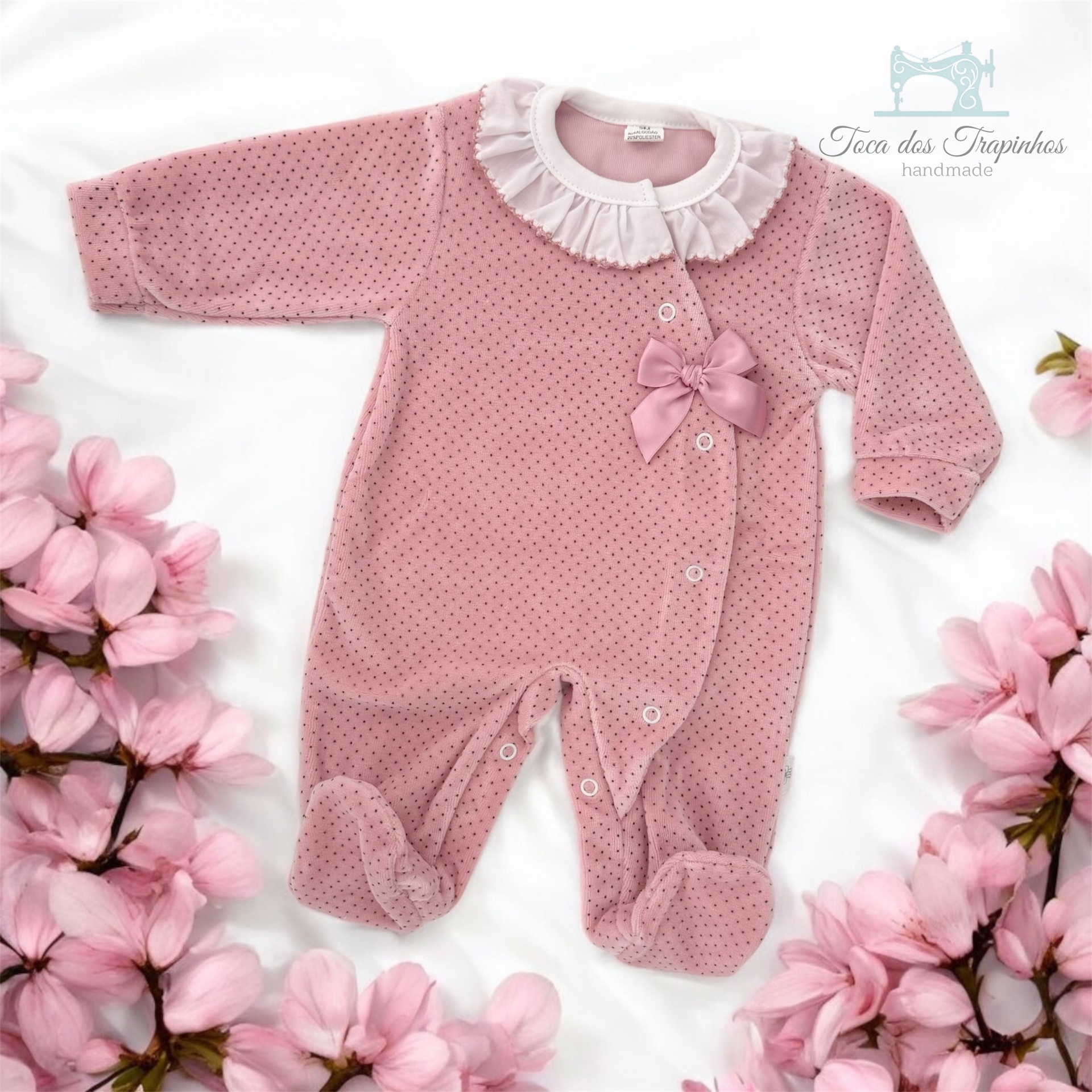 Macacão rosa para bebé com gola branca e pequeno laço rosa, rodeado por flores cor-de-rosa