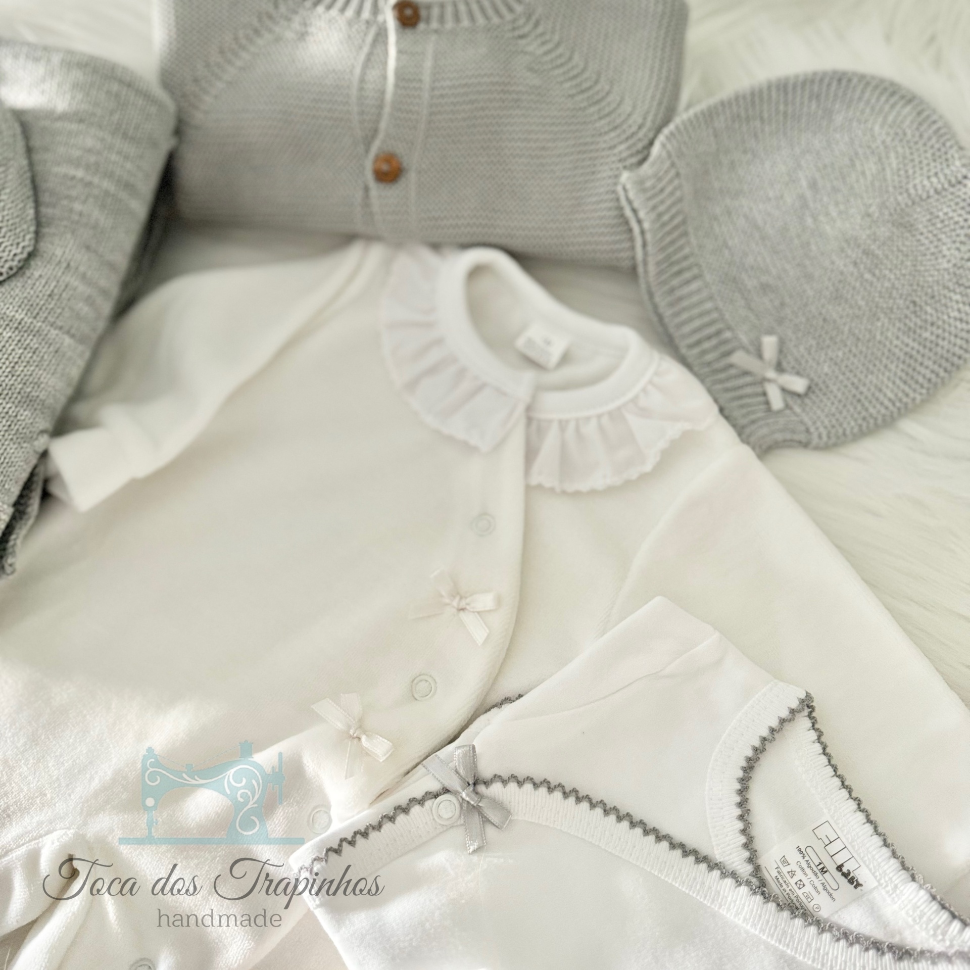 Roupas de bebé em branco e cinzento, incluindo casaco com babados e gorro com laço