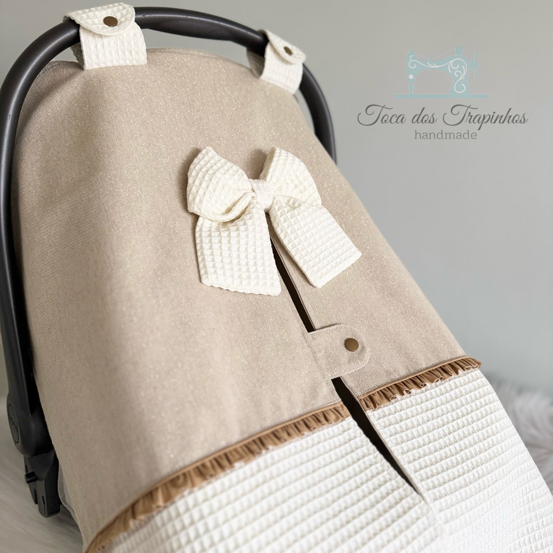 Capa para carrinho de bebé castanha com laço branco e botões de pressão, fundo cinzento com logo Toca dos Trapinhos handmade