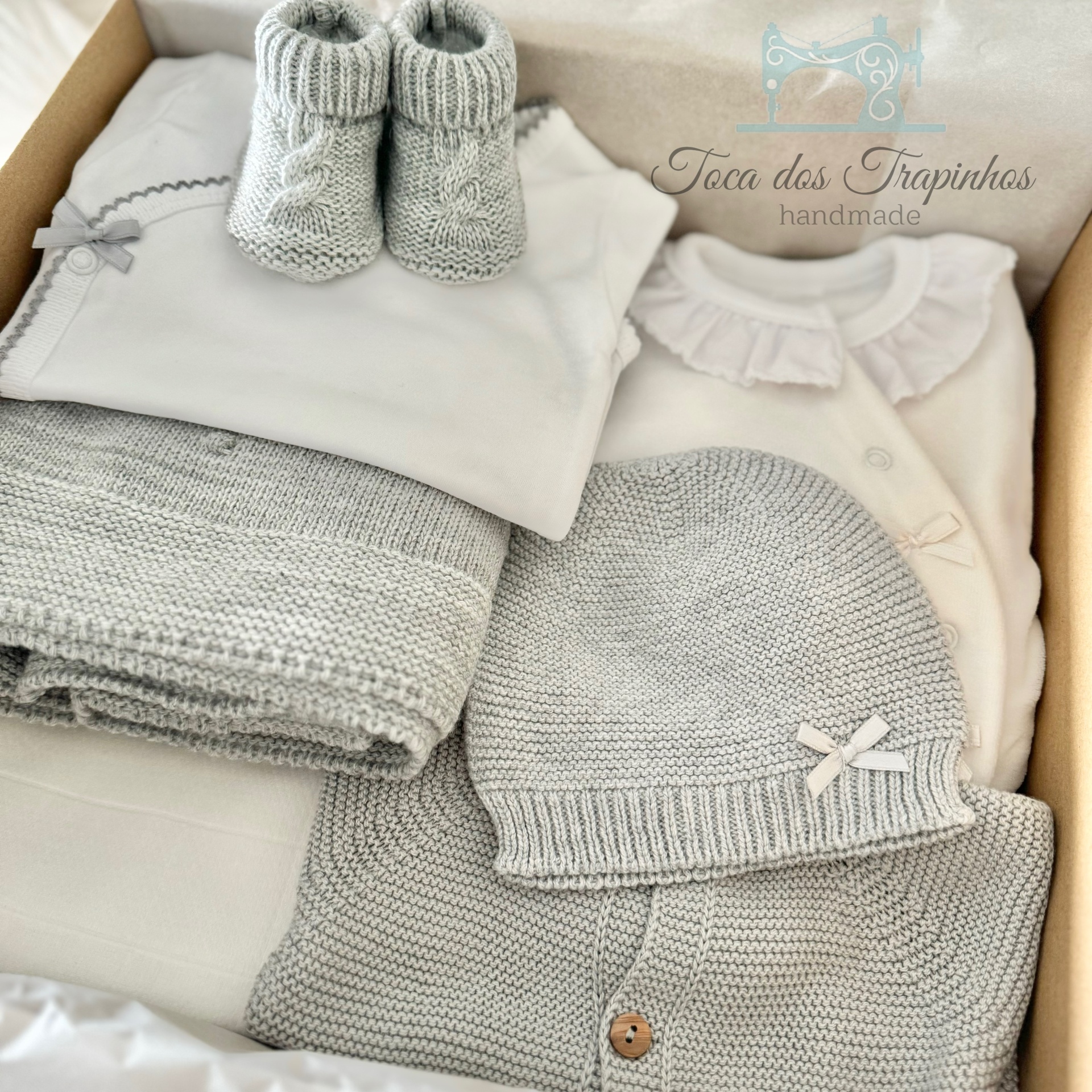 Conjunto de roupa para bebé cinza e branca com malha e algodão, em caixa castanha clara.