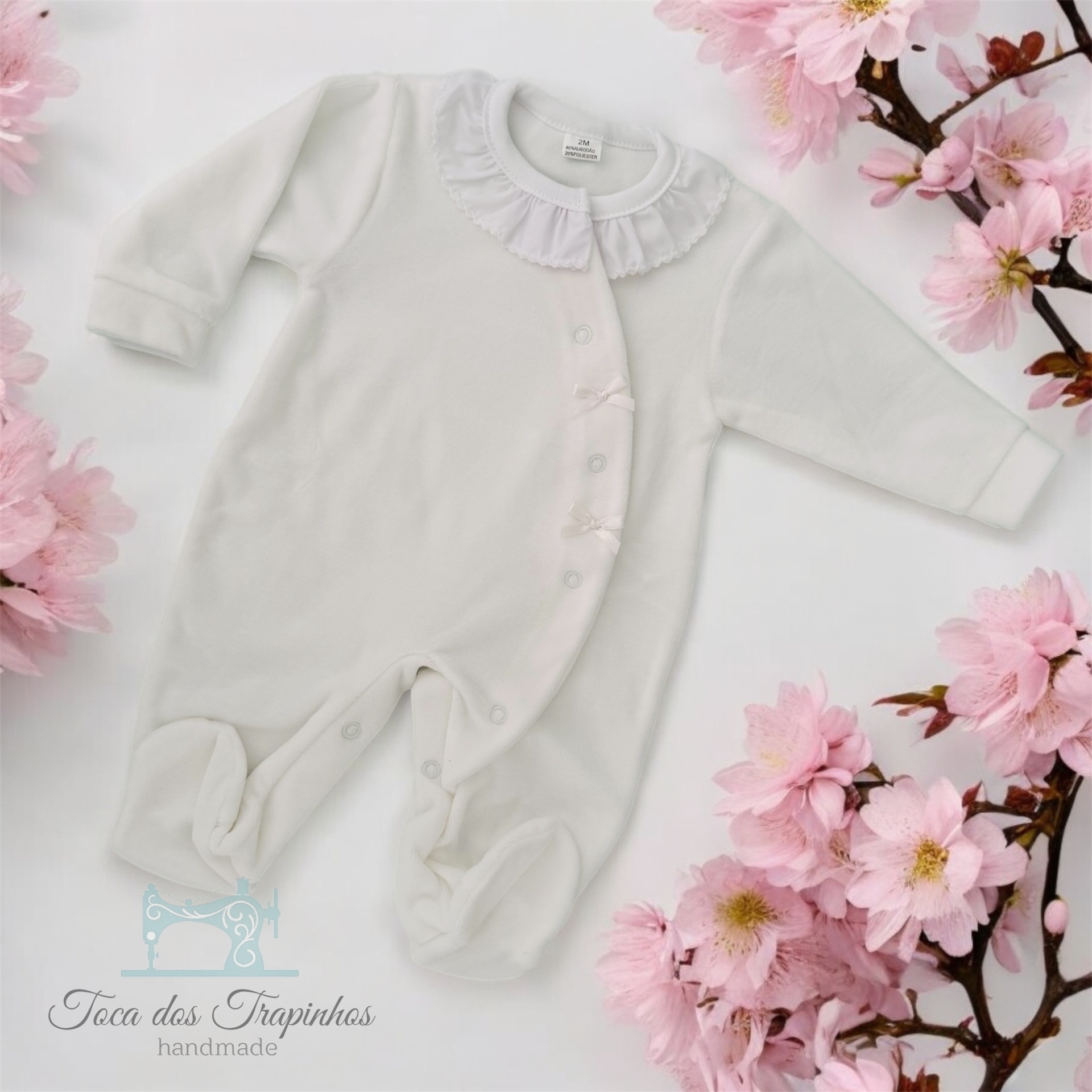 Macacão branco de bebé com gola franzida e botões, rodeado por flores cor-de-rosa