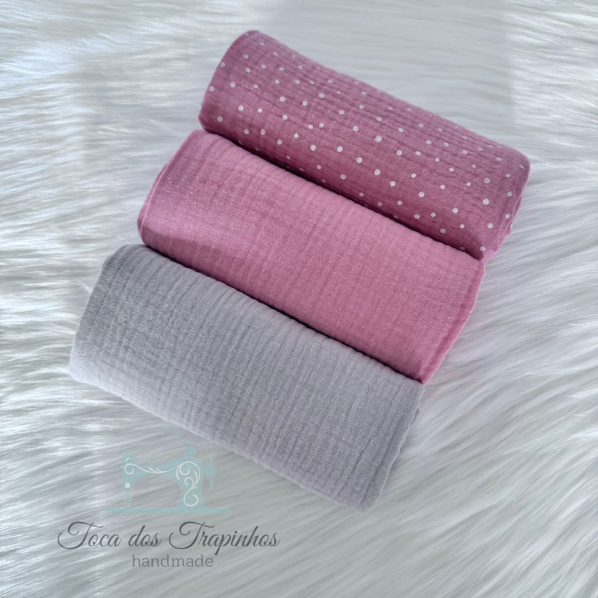 Três mantas macias em tons de rosa e cinza sobre superfície branca felpuda.