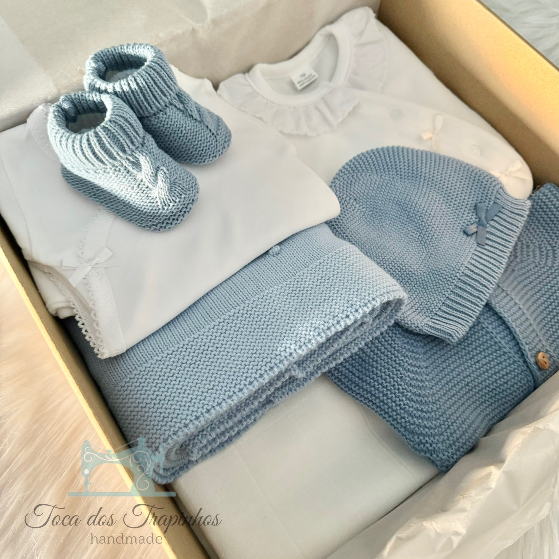 Conjunto de roupa de bebê azul claro e branco com sapatinhos, touca, blusa e manta de malha dentro de caixa de madeira