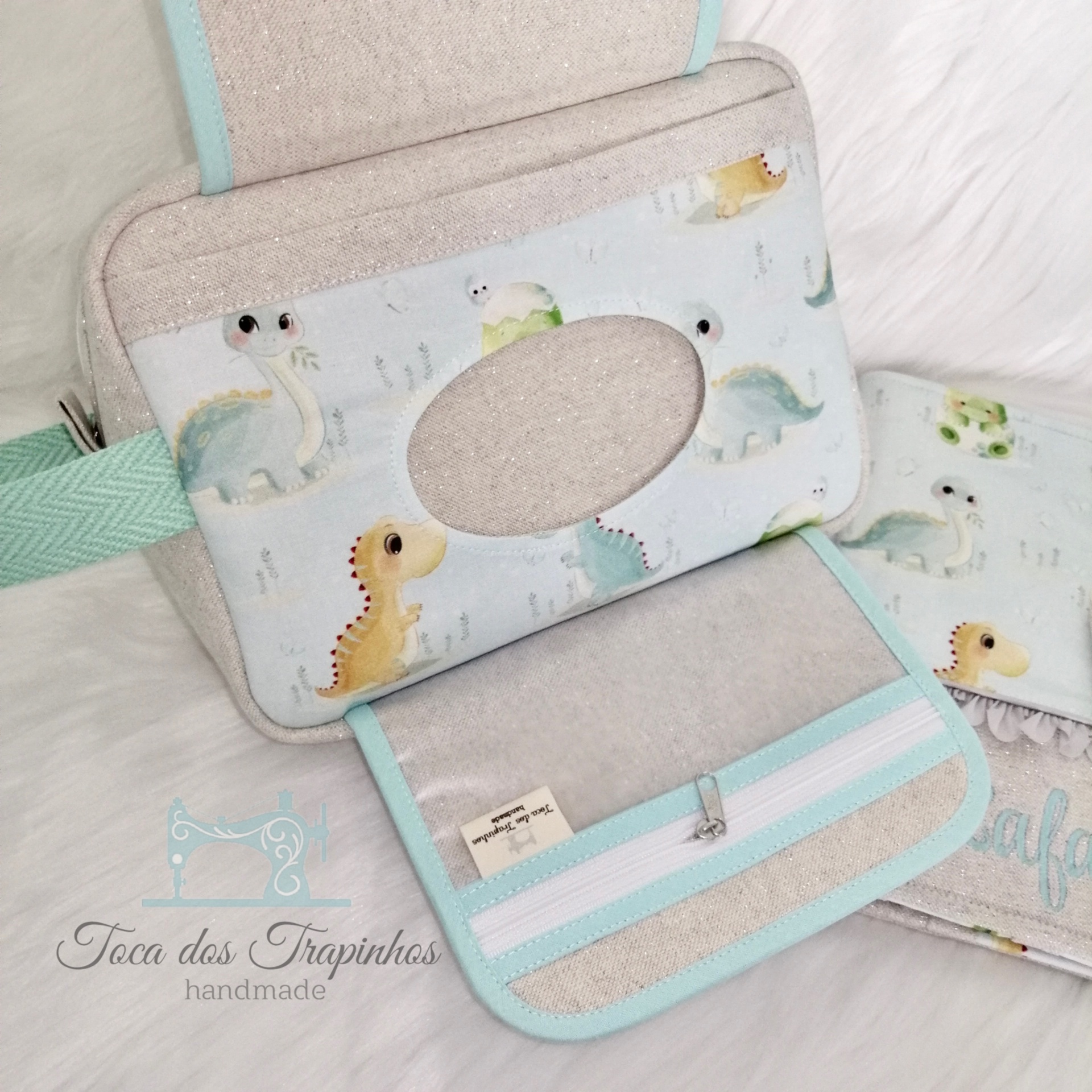 Bolsa ou estojo azul claro com dinossauros, aba aberta mostrando bolso interior com fecho