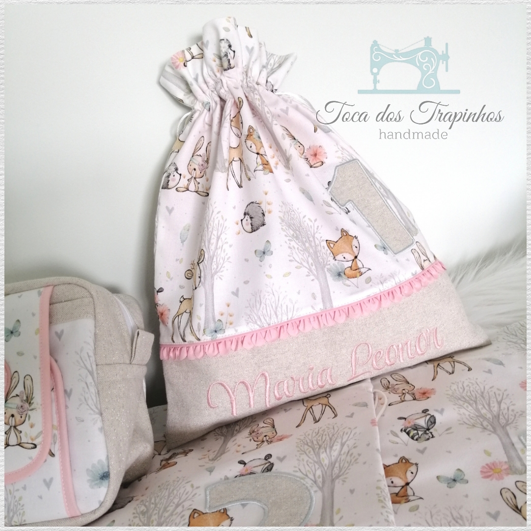Saco de tecido com estampado de animais da floresta e nome bordado, com mala e roupa de cama ao fundo