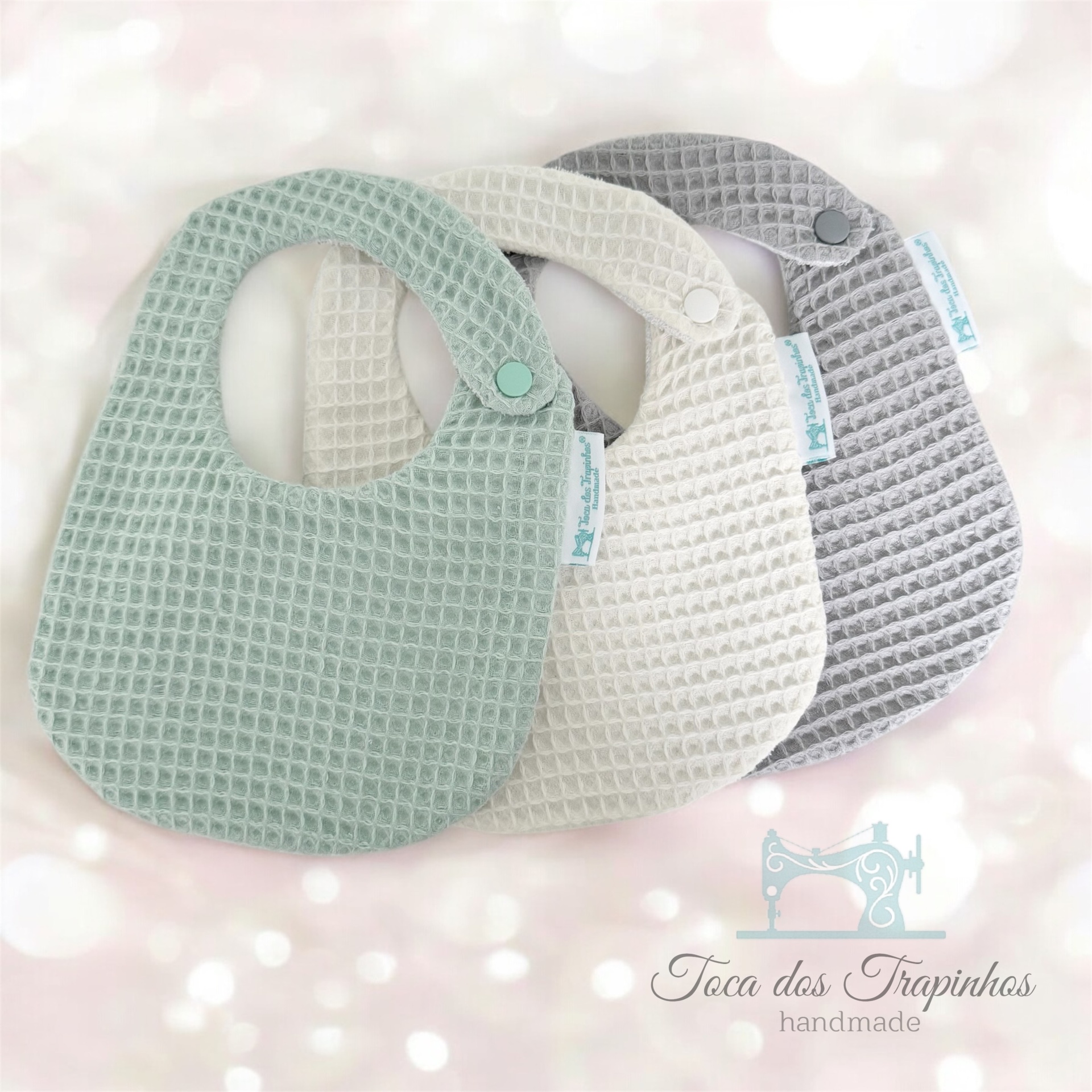 Três babetes de tecido com padrão waffle em verde claro, bege e cinza com fecho de pressão