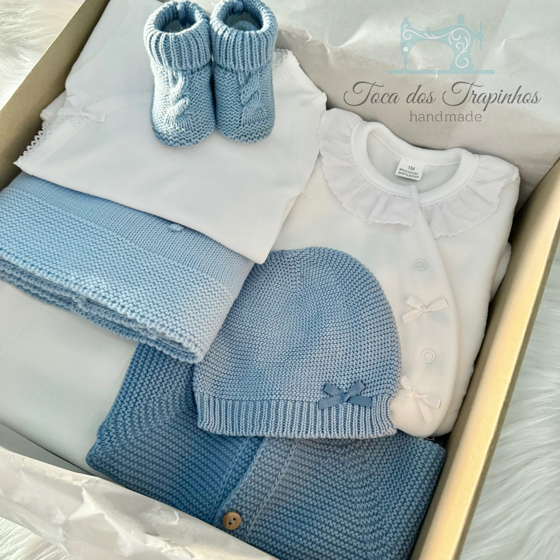 Conjunto de roupa de bebé azul e branco com gorro, botins, casaco e body numa caixa com papel branco