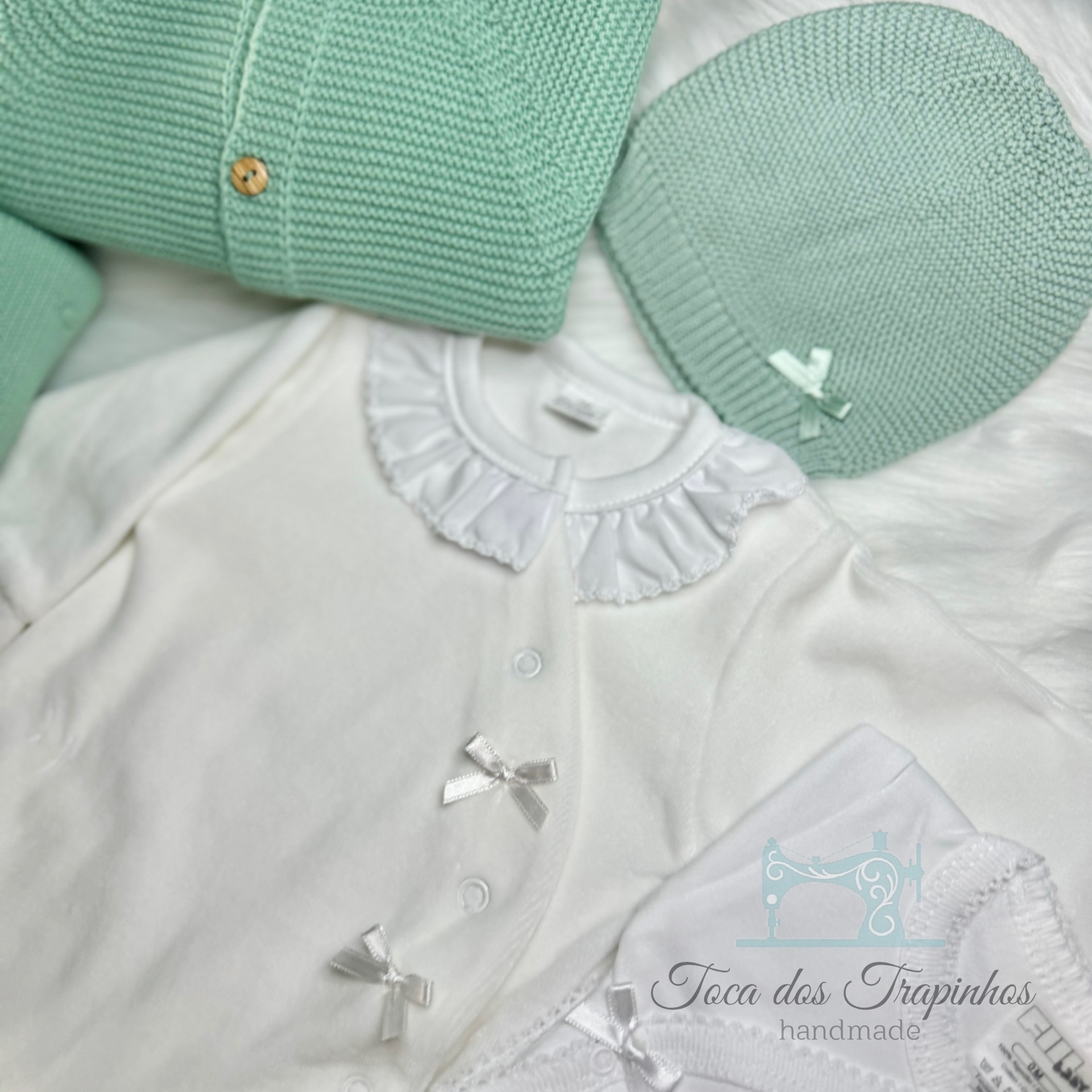 Conjunto de roupa de bebé branco e verde água com gola de folho, laços e manta em tecido felpudo branco.