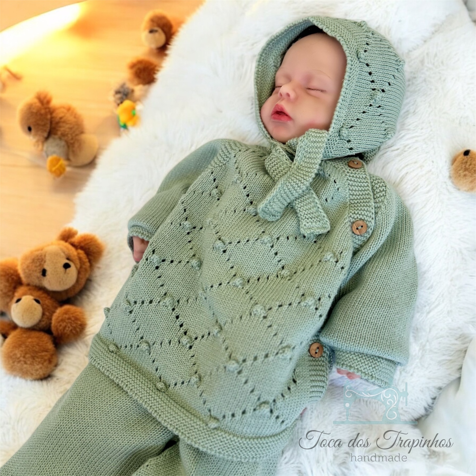 bebé vestido com conjunto de malha verde claro com capuz e poncho, deitado numa manta branca com ursos de peluche ao fundo