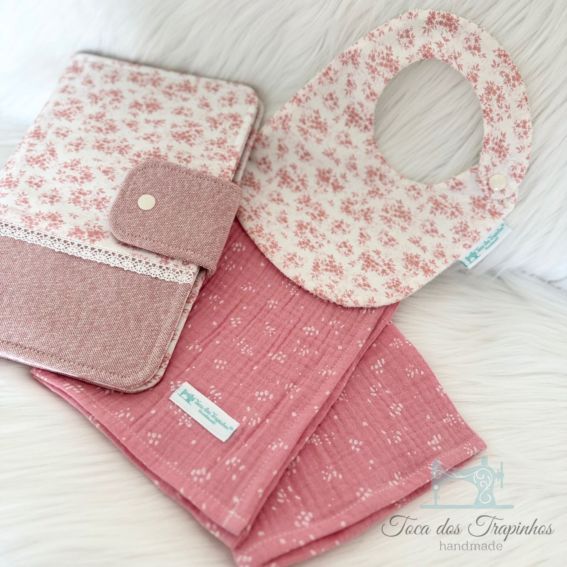 Conjunto de babete, pano e bolsinha, em rosa e bege, sobre fundo branco felpudo