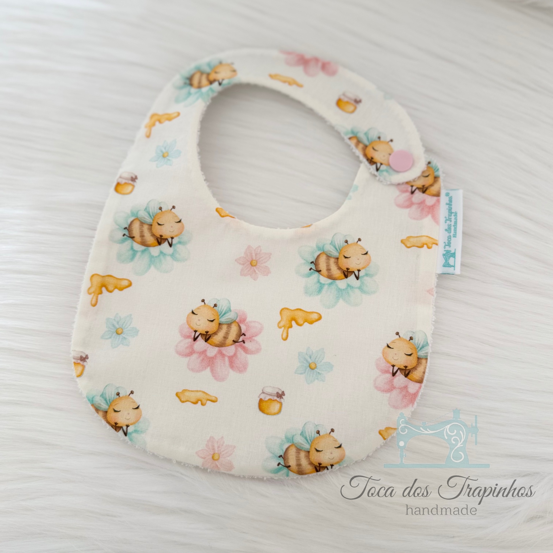 Babete branco com padrão de abelhas amarelas sobre flores coloridas e fecho rosa