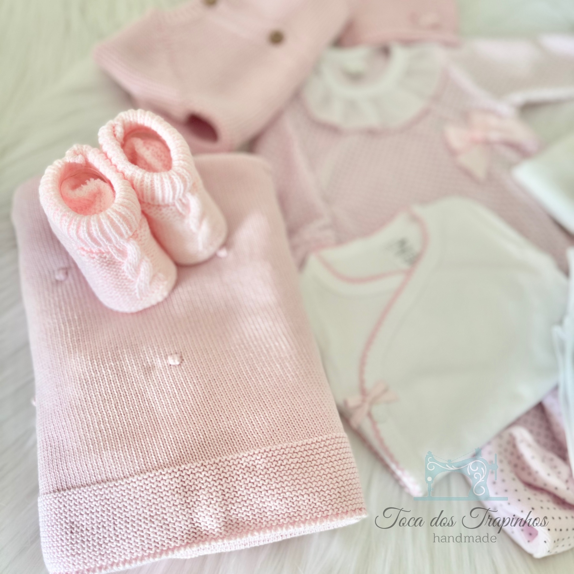 Conjunto de roupa de bebé cor-de-rosa com manta, botinhas e body sobre superfície branca felpuda