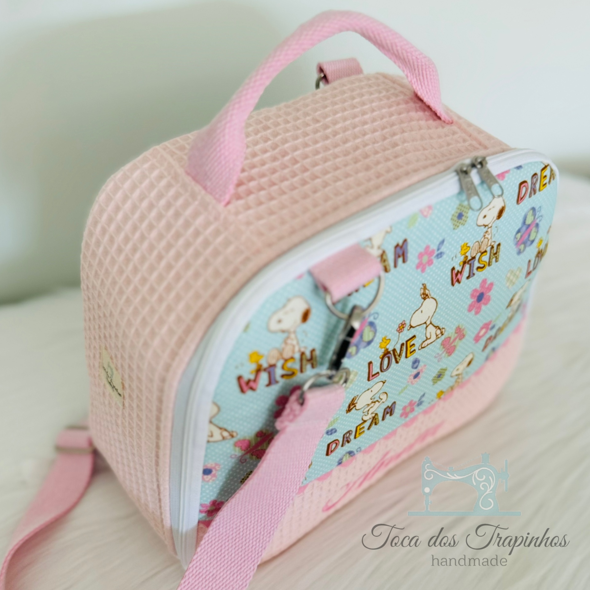 Mala infantil rosa e azul claro com desenhos e palavras em inglês, alças rosa, fecho metálico, colocada sobre superfície branca