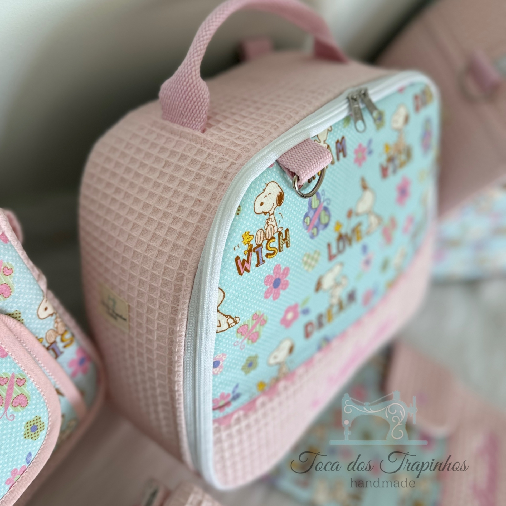 Bolsa infantil rosa com painel azul claro e padrão Snoopy, com texto e fecho branco